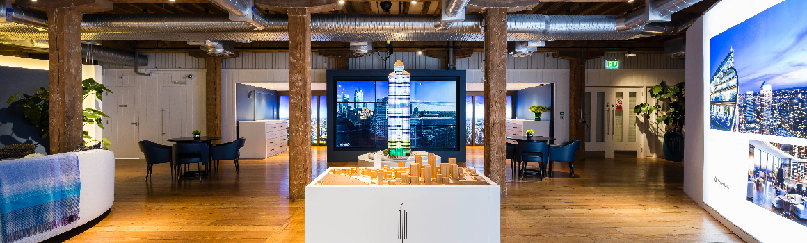 A modern showroom features a detailed architectural model of a skyscraper on a central pedestal. The room has wooden floors, exposed ceiling pipes, and large wall art depicting urban landscapes illuminated by LED backlit graphics. Several seating areas and potted plants create an inviting atmosphere.