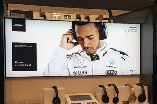 Advertisement featuring F1 driver Louis Hamilton with sponsor logos. The banner, illuminated by Bright Green Technology LED backlit graphics, reads "Focus comes first" and showcases various headphones displayed below the image.