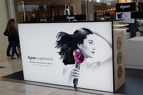 A Bright Green Technology LED lightbox displays an advertisement featuring a woman using a Dyson Supersonic hairdryer. The kiosk is decorated with the product image and description highlighting "intelligent heat control for shine" and is situated in a busy shopping area.