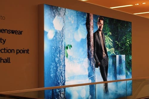 A large backlit advertisement displays a man in a dark suit leaning against a tree in a forest setting. The advertisement is placed on the wall of a modern indoor space.
