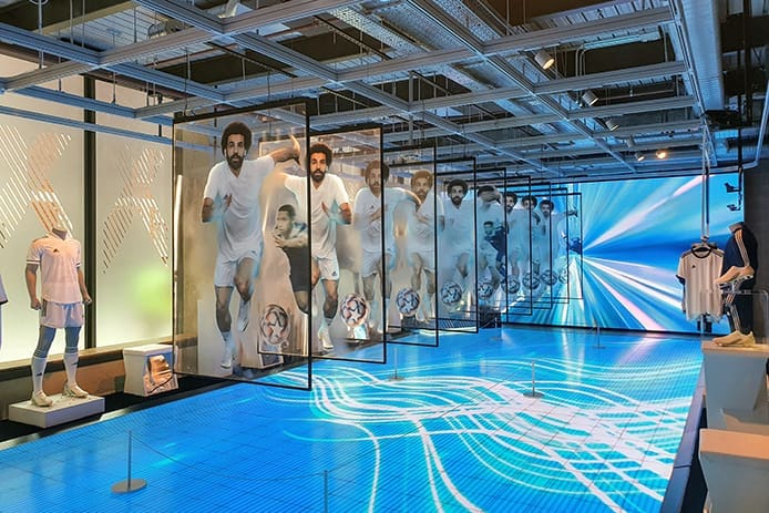 A futuristic store display featuring multiple life-sized screens displaying footballer Mohamed Salah. The floor and background are illuminated with dynamic blue and white light patterns, enhanced by LED light panels.