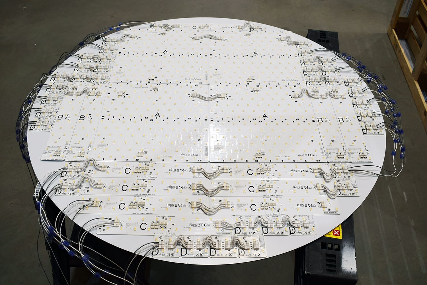 A large, round circuit board mounted on a table. It is populated with numerous LED lights and interconnected wiring. The board has labeled sections marked 'A', 'B', 'C', and 'D', and the setup appears to be part of an electronic assembly or testing process.