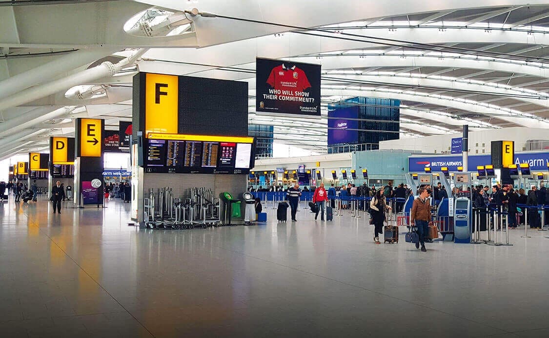 Heathrow airport terminal with tall ceilings and large windows. Passengers are walking and standing in line at check-in counters. Overhead signage indicates gates illuminated by LED grids.