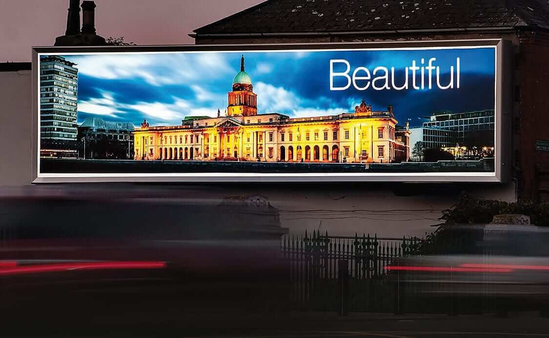 A large outdoor billboard displays a nighttime photo of an illuminated, historic building with a clock tower, under a dramatic cloudy sky. Enhanced by LED backlit graphics, the scene is set against urban buildings and moving traffic.