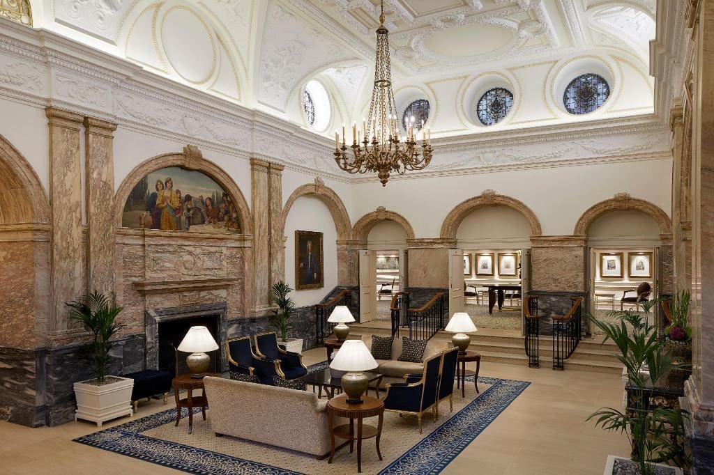 A luxurious, spacious lobby with a high, ornate ceiling, arched windows, and chandeliers. It features an elegant seating area with sofas and chairs around coffee tables, surrounded by tall columns, plants, classical artwork on the walls, and backlit glass with tuneable white LEDs.