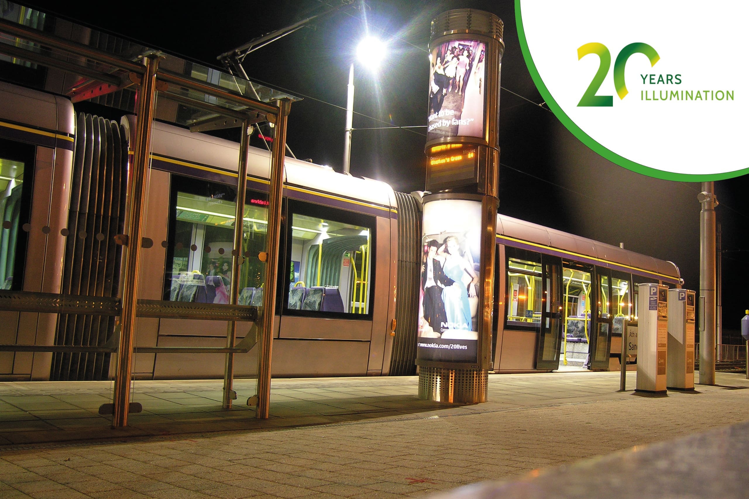 A tram is stopped at a well-lit station at night, where digital screens with LED backlit graphics display advertisements. A logo in the upper right corner reads "20 Years of Illumination" in green text.