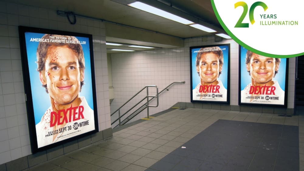 The New York City Underground subway station boasts tiled walls adorned with LED backlit posters for the TV show "Dexter." Each poster showcases a man smiling, his face marked with blood splatters, and a "20 Years Illumination" logo in the top right corner.