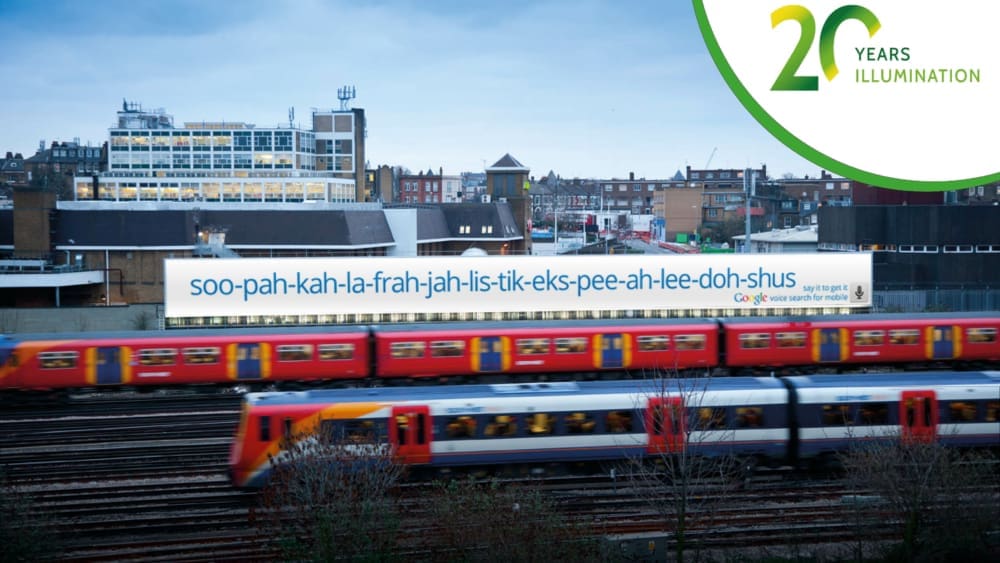 A train moves swiftly on tracks in an urban area, with buildings and houses in the background. An illuminated billboard featuring large format advertising displays the word "soo-pah-kah-la-frah-jah-lis-tik-eks-pee-ah-lee-doh-shus." A "20 Years Illumination" logo is in the top-right corner.