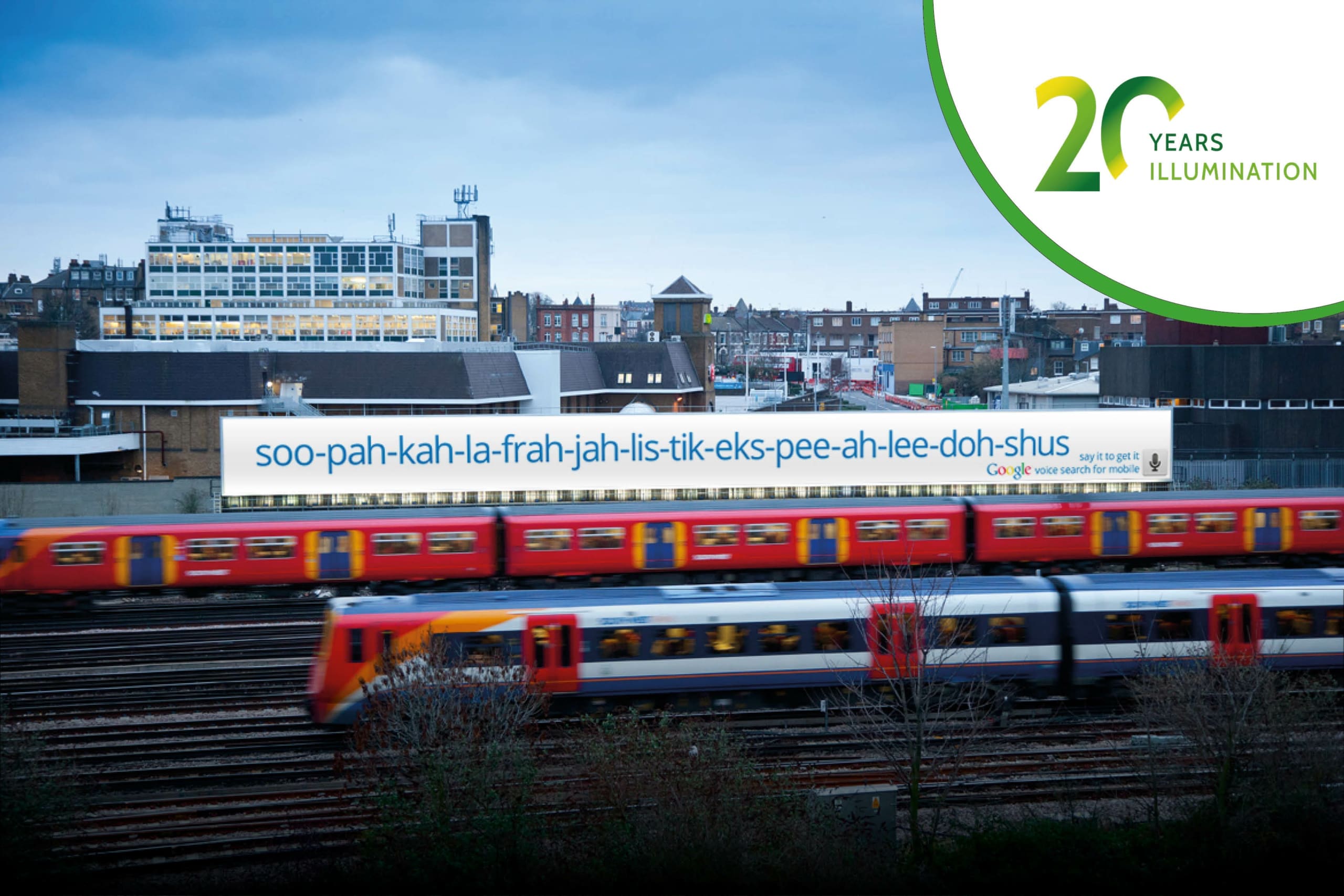 A train moves swiftly on tracks in an urban area, with buildings and houses in the background. An illuminated billboard featuring large format advertising displays the word "soo-pah-kah-la-frah-jah-lis-tik-eks-pee-ah-lee-doh-shus." A "20 Years Illumination" logo is in the top-right corner.