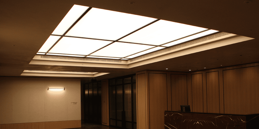 A modern interior space featuring large, LED light panels in the ceiling. The walls are adorned with light wood paneling, complemented by recessed lighting fixtures. A glass partition can be seen on the left side, and the area is minimally furnished.