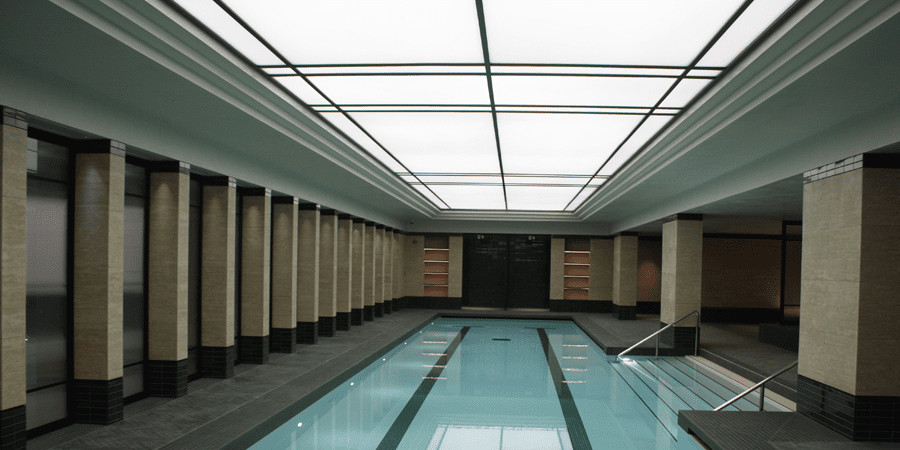A modern indoor swimming pool with a sleek, minimalist design. The pool is surrounded by beige tiled walls with dark trim and lightly frosted windows. The ceiling features illuminated light panels. There are steps and a metal railing leading into the pool.