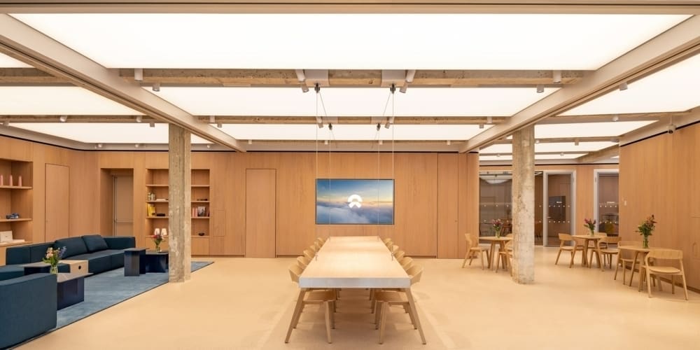 A modern, well-lit office space featuring a large conference table surrounded by wooden chairs. The LED illuminated ceiling adds to the ambiance, highlighting a flat-screen TV displaying a scenic image mounted on the back wall.