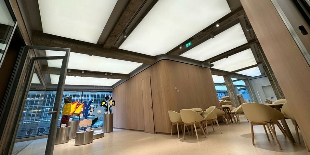 An open and modern cafeteria with wooden furnishings and an LED light panel brightly illuminating the ceiling. To the left, colorful abstract sculptures are displayed near large windows that offer a view of a cityscape. Beige chairs surround wooden tables on a light-colored floor.