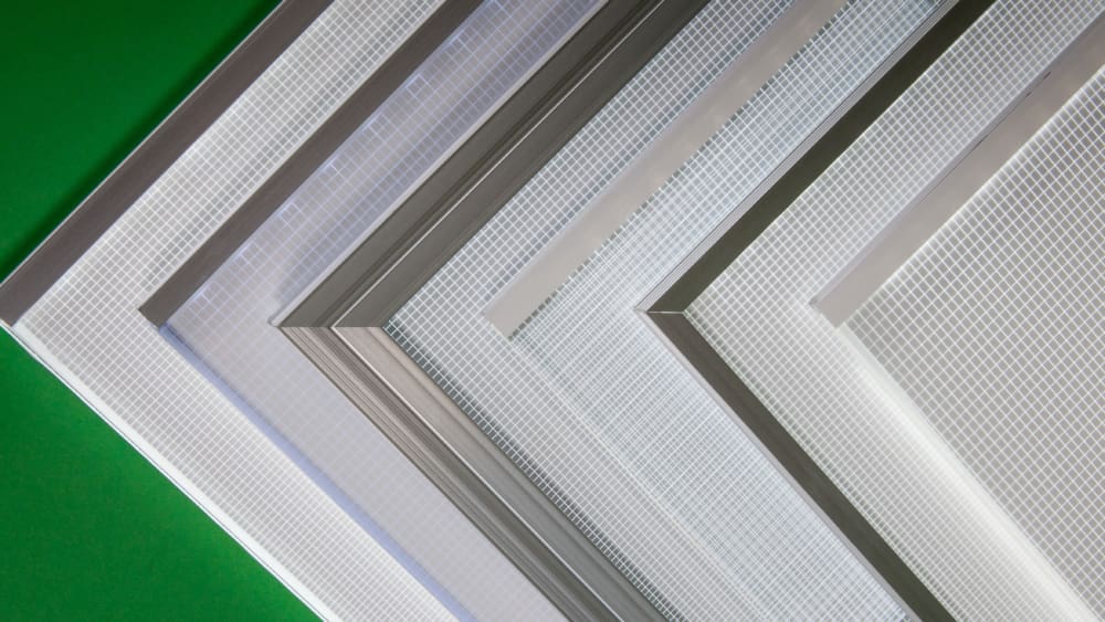 A diagonal arrangement of multiple window screens with varying mesh sizes and frame materials set against a green background features an integrated LED Light Sheet. The screens overlap slightly, showing a comparison of their different build attributes.