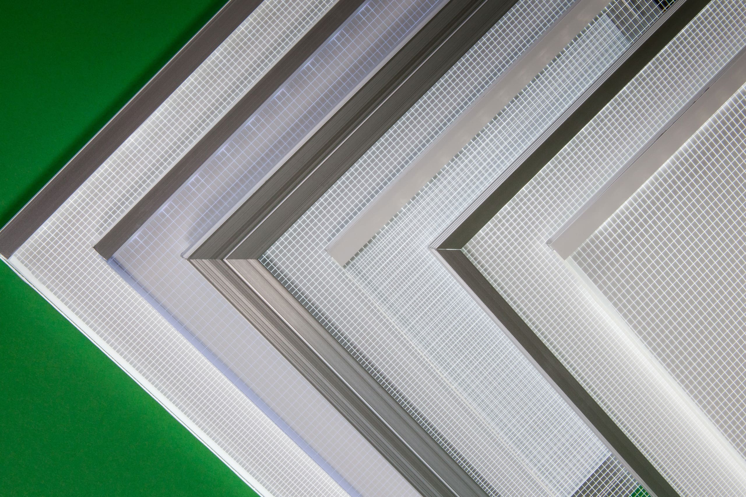 A diagonal arrangement of multiple window screens with varying mesh sizes and frame materials set against a green background features an integrated LED Light Sheet. The screens overlap slightly, showing a comparison of their different build attributes.